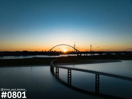 Luchtfoto van De Oversteek over de Waal bij Nijmegen, met de zon onder de boog en een lage brug op pijlers op de voorgrond