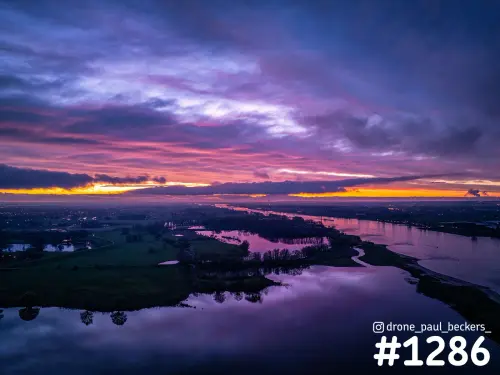 Luchtfoto van een rivierlandschap bij Nijmegen na zonsondergang, met paarse wolken en oranje gloed boven spiegelend water