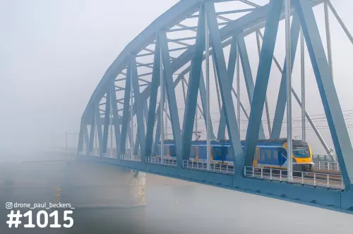 Luchtfoto van een stalen boogbrug in dichte mist, met een gele en blauwe trein tussen het blauwe vakwerk