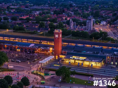Luchtfoto van station Nijmegen met de rode klokkentoren in het blauwe uur, de verlichte hal en fietsenstalling op de voorgrond