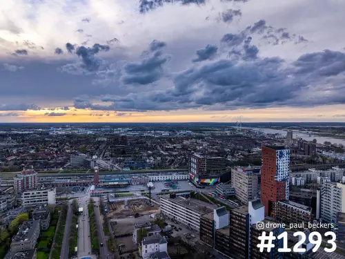 Luchtfoto van het Stationsgebied in Nijmegen met rode hoogbouw, donkere wolken en oranje schemerlicht boven de Waal