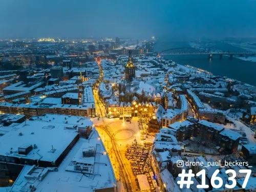 Luchtfoto van de Stevenskerk en de Grote Markt in Nijmegen in de sneeuw, met verlichte daken en de Waal op de achtergrond