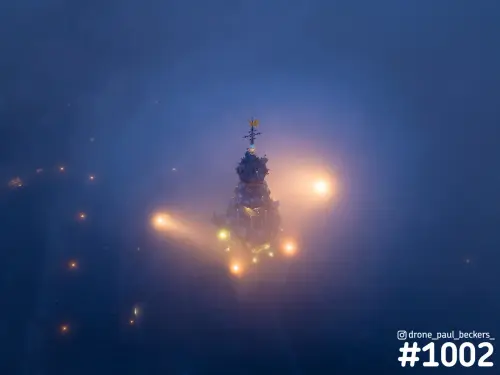 Luchtfoto van de torenspits van de Stevenskerk in Nijmegen in dichte mist, met warme lichten die door de avondschemering gloeien