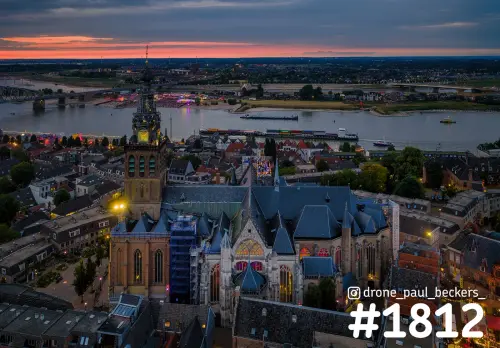 Luchtfoto van de Stevenskerk in Nijmegen in de avondschemering, met steigers, verlichte toren en de Waal erachter