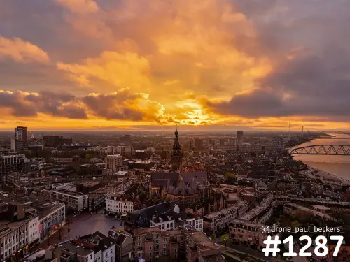 Luchtfoto van de Stevenskerk en het centrum van Nijmegen onder oranje wolken, met de Waal en een brug aan de horizon