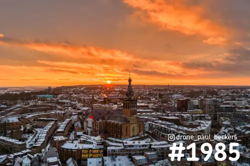 Luchtfoto van de Stevenskerk in het centrum van Nijmegen bij zonsondergang, met sneeuw op de daken en een oranje lucht