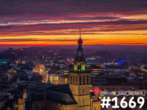Luchtfoto van de Stevenskerk in Nijmegen na zonsondergang, met groen verlichte toren en een oranjerode lucht boven het centrum