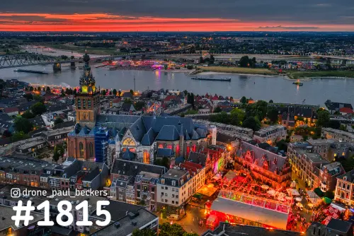 Luchtfoto van de Stevenskerk in Nijmegen bij schemering, met verlichte kermis op de Grote Markt en de Waal met rode lucht erachter