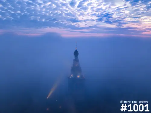 Luchtfoto van de Stevenskerk in Nijmegen die uit dikke mist oprijst, met een lichtstraal en roze avondwolken