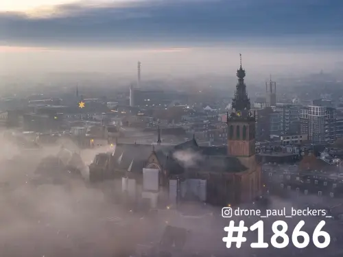 Luchtfoto van de Stevenskerk in Nijmegen met laaghangende mist tussen de daken en een verlichte ster in de verte