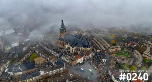 Luchtfoto van de Stevenskerk in Nijmegen met laaghangende mist boven de daken en het plein, herfstkleuren in de bomen
