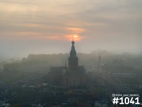 Luchtfoto van de Stevenskerk in Nijmegen in dichte mist, met oranje zon die opkomt achter de toren en een reuzenrad ernaast