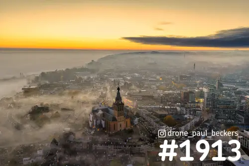 Luchtfoto van de Stevenskerk in het centrum van Nijmegen met mist tussen de daken en een oranje gloed aan de horizon
