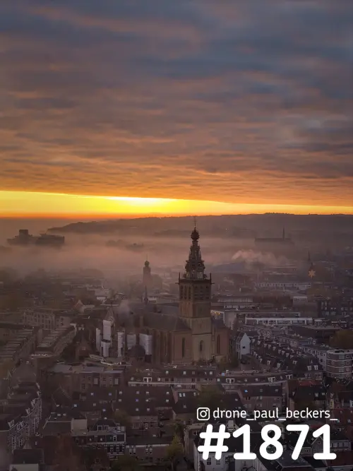Luchtfoto van de Stevenskerk in het centrum van Nijmegen bij zonsopkomst, met mist tussen de daken en een oranje lucht