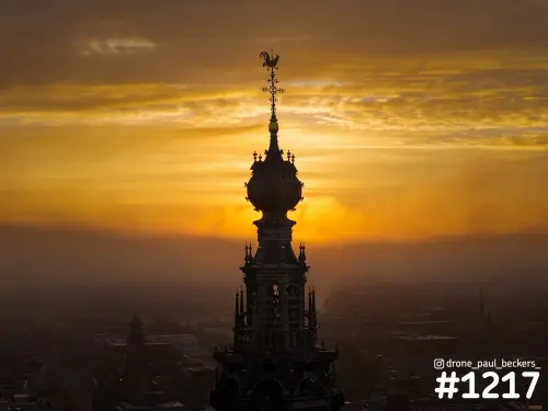 Luchtfoto van de torenspits van de Stevenskerk in Nijmegen als silhouet tegen een oranje zonsopkomst, met mist boven het centrum