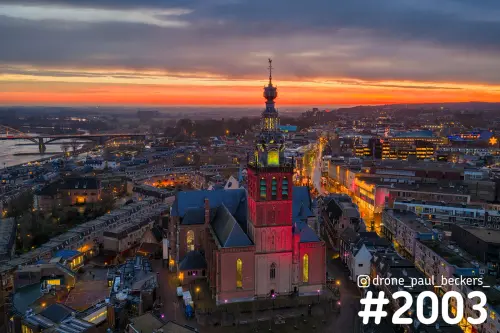 Luchtfoto van de verlichte Stevenskerk in Nijmegen in de schemering, met oranje gloed boven de Waal en het centrum