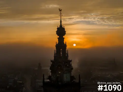 Luchtfoto van de Stevenskerk in Nijmegen als silhouet tegen een oranje lucht, met de zon laag boven mistige daken