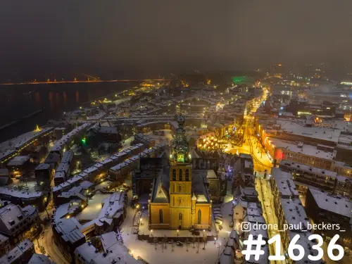 Luchtfoto van de Stevenskerk in Nijmegen in de sneeuw bij avond, met verlichte daken, de Grote Markt en de Waal erachter