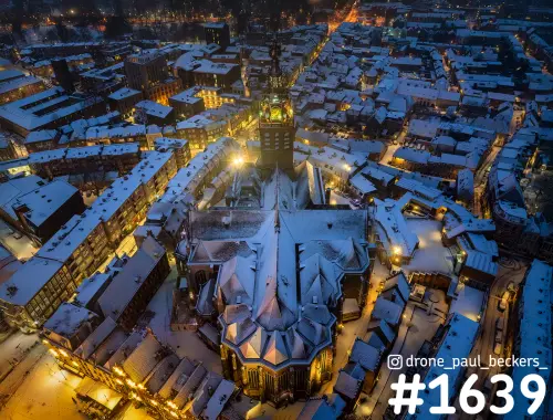 Luchtfoto van de Stevenskerk in Nijmegen in de sneeuw bij blauwe schemering, met verlichte toren en besneeuwde daken
