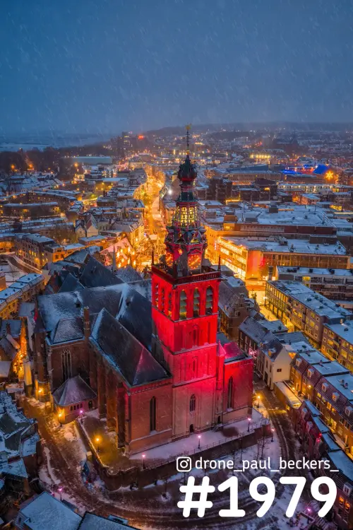 Luchtfoto van de rood verlichte Stevenskerk in Nijmegen in de avond, met sneeuwval en besneeuwde daken in het centrum