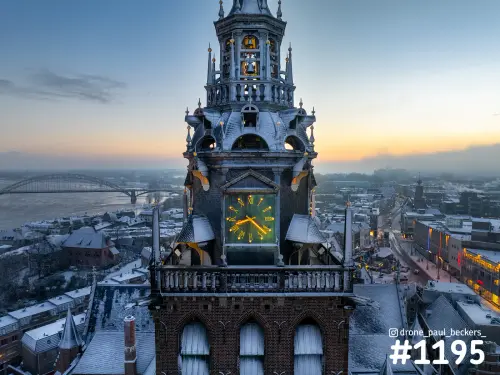 Luchtfoto van de torenspits van de Stevenskerk in Nijmegen in de schemering, met besneeuwde daken en de Waalbrug op de achtergrond