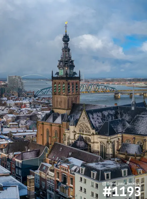 Luchtfoto van de Stevenskerk in Nijmegen met sneeuw op de daken, de Waal en de boog van de Oversteek op de achtergrond