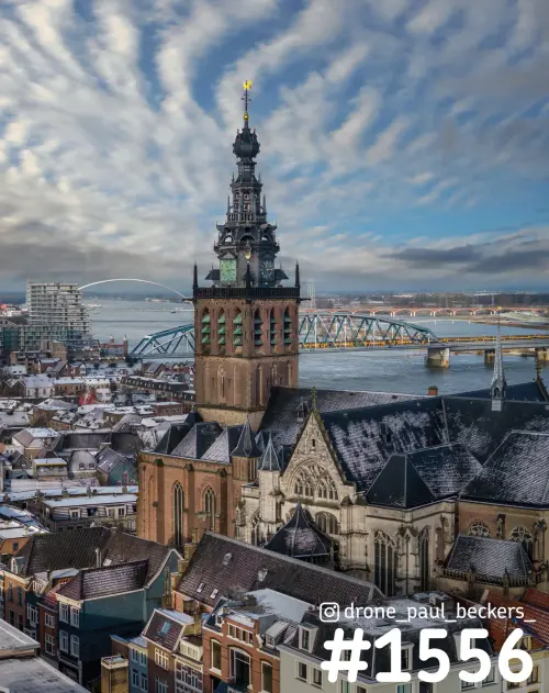 Luchtfoto van de Stevenskerk in Nijmegen met sneeuw op de daken, de Waal en de Oversteek onder een gestreepte wolkenlucht