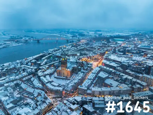 Luchtfoto van de Stevenskerk en besneeuwde daken in Nijmegen, met de Waalbrug in de verte in blauw avondlicht