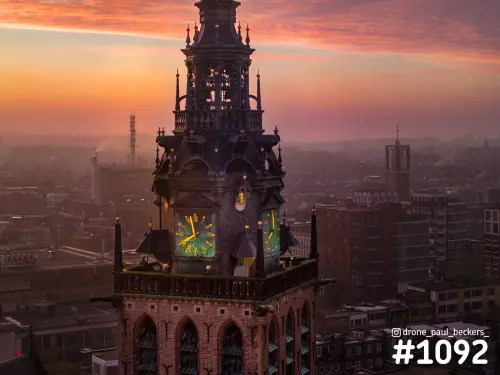 Luchtfoto van de Stevenskerk in Nijmegen: toren met groen verlichte wijzerplaten, roze lucht en nevel boven de daken