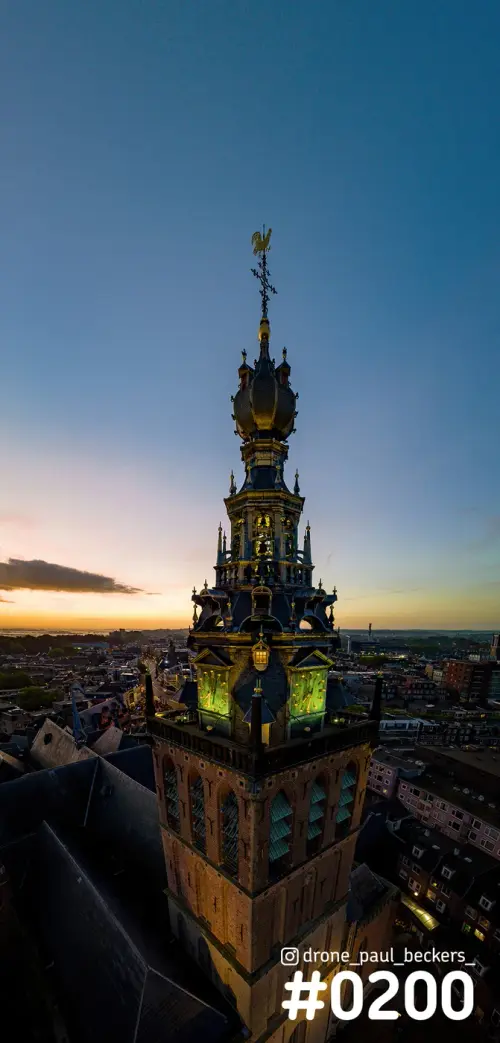 Luchtfoto van de toren van de Stevenskerk in Nijmegen bij schemering, met verlichte klokkenwijzers en een oranje gloed aan de horizon