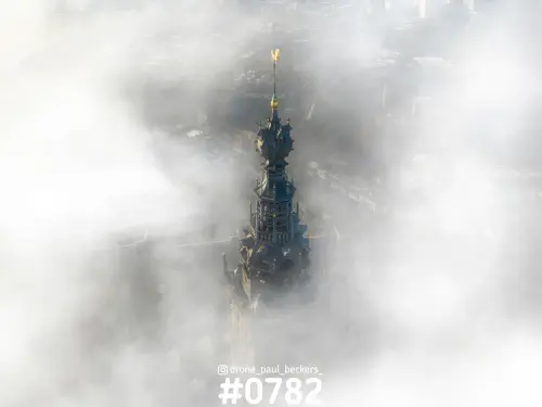 Luchtfoto van de Stevenskerk in Nijmegen: de torenspits met gouden haan steekt door een dik mistdek heen