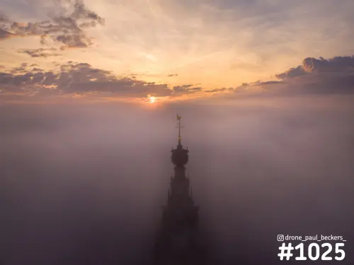 Luchtfoto van de torenspits van de Stevenskerk in Nijmegen die boven dichte mist uitsteekt, met zonsopkomst achter de wolken