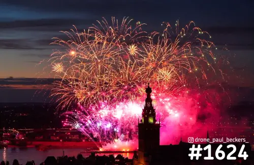 Luchtfoto van vuurwerk boven de Stevenskerk in Nijmegen in de avondschemering, met oranje en roze lichtsporen