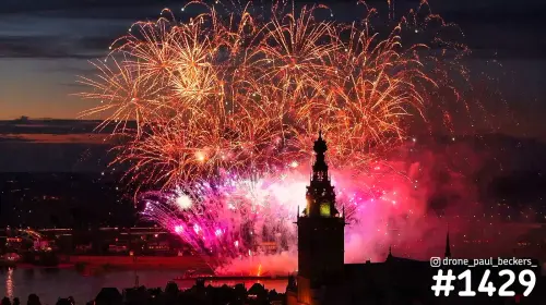 Luchtfoto van vuurwerk boven de Stevenskerk en de Waal in Nijmegen, met oranje explosies en roze rook bij schemering
