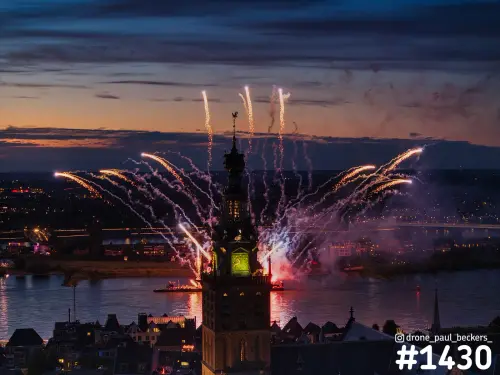Luchtfoto van vuurwerk boven de Waal in Nijmegen, gezien langs de Stevenskerktoren in het avondlicht, met Lent op de achtergrond