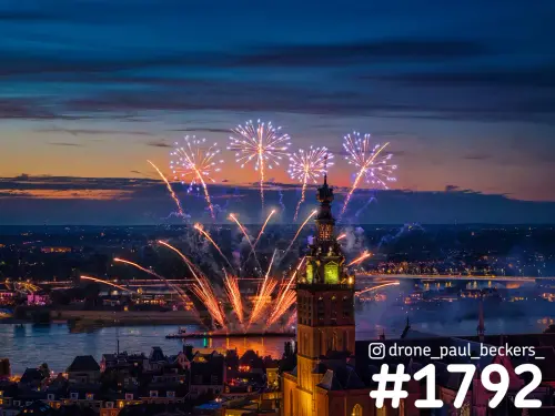 Luchtfoto van vuurwerk boven de Waal in Nijmegen, met de verlichte toren van de Stevenskerk en Lent aan de overkant