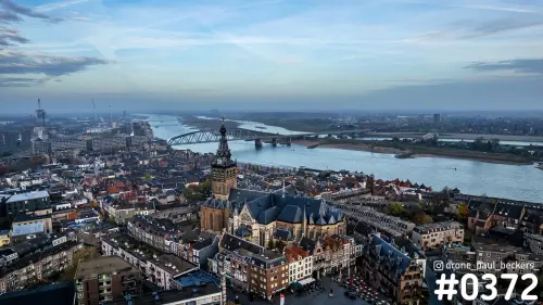 Luchtfoto van de Stevenskerk in Nijmegen met de Waal en een stalen boogbrug in de verte, onder heiige herfstlucht
