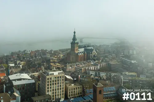 Luchtfoto van de Stevenskerk in Nijmegen in de mist, met de Waal en een brug op de achtergrond boven de daken