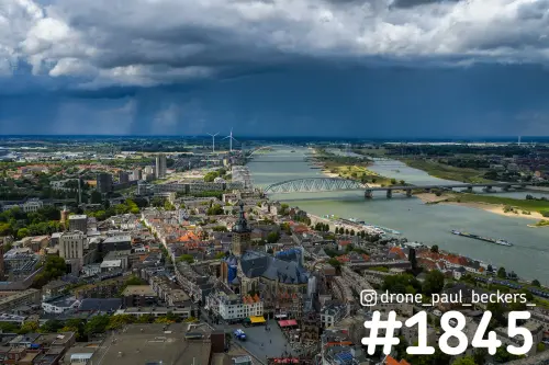 Luchtfoto van Nijmegen met Stevenskerk, de Waal en De Oversteek onder donkere regenwolken en zonlicht op de daken