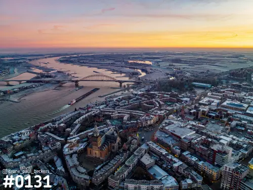 Luchtfoto van het centrum van Nijmegen met de Stevenskerk, de Waal en de Waalbrug onder een oranje lucht bij schemering