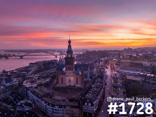 Luchtfoto van de Stevenskerk in het centrum van Nijmegen met de Waal en Waalbrug onder een roze en oranje schemerlucht