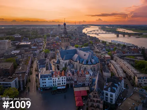 Luchtfoto van de Stevenskerk in Nijmegen bij zonsondergang, met de Waal en een stalen brug onder een oranje lucht