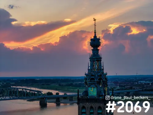 Luchtfoto van de toren van de Stevenskerk in Nijmegen bij zonsondergang, met oranje wolken boven de Waal en Lent