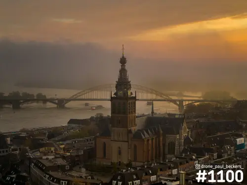 Luchtfoto van de Stevenskerk in Nijmegen met de Waalbrug in de mist en een gouden lucht boven de Waal