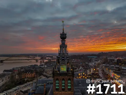 Luchtfoto van de Stevenskerktoren in Nijmegen onder een oranje lucht, met de Waalbrug en de Waal op de achtergrond
