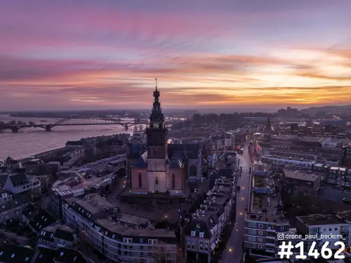 Luchtfoto van de Stevenskerk in Nijmegen bij schemering, met de Waalbrug over de Waal en een roze-oranje lucht