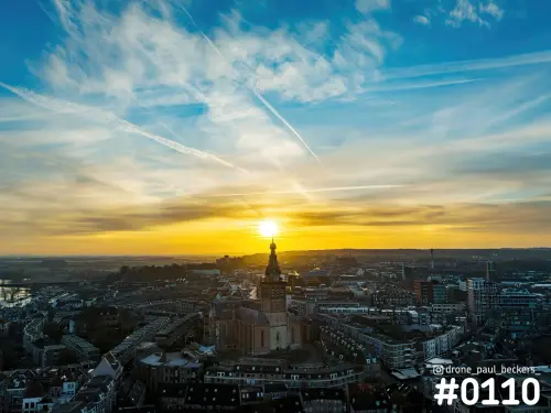 Luchtfoto van de Stevenskerk in Nijmegen bij zonsondergang, met de zon aan de horizon en gestreepte wolken erboven
