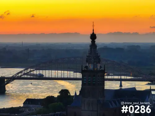 Luchtfoto van de Stevenskerk in Nijmegen met de Waalbrug erachter, oranje schemerlucht en gouden weerkaatsing op de Waal
