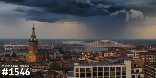 Luchtfoto van de Stevenskerk en de Waalbrug in Nijmegen in de avondschemering, met een donkere regenbui boven de rivier