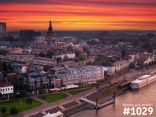 Luchtfoto van de Stevenskerk en de Waalkade in Nijmegen onder een oranjerode avondlucht, met een rivierschip op de Waal
